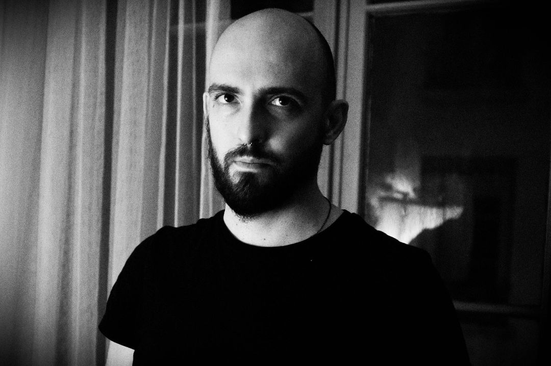 potrait noir et blanc de l'auteur regardant l'objectif, posant devant la fenêtre d'un appartement et portant un t-shirt noir